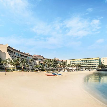 Al Raha Beach Resort & Spa Abu Dhabi Eksteriør bilde The photo depicts a serene beach scene with a clear blue sky. In the foreground, there is a sandy beach that gently slopes down to calm water. On the left side, several palm trees provide a tropical atmosphere. In the background, there are two large 