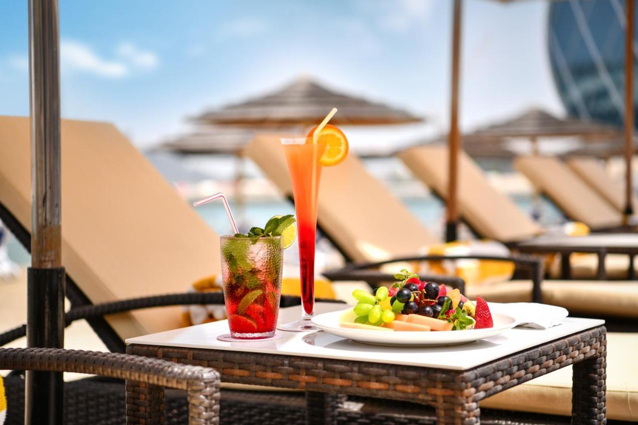 Al Raha Beach Resort & Spa Abu Dhabi Eksteriør bilde The photo shows a table set outdoors, featuring two colorful cocktails and a plate of food. One cocktail is a vibrant red drink garnished with mint, while the other is a layered orange beverage with an orange slice on the rim. The plate of food inclu