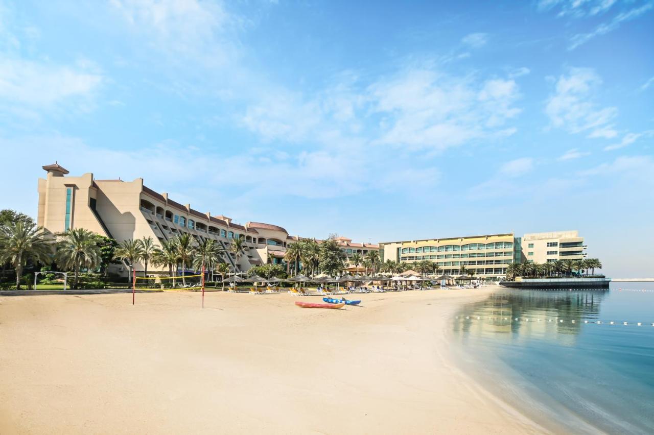 Al Raha Beach Resort & Spa Abu Dhabi Eksteriør bilde The photo depicts a serene beach scene with a clear blue sky. In the foreground, there is a sandy beach that gently slopes down to calm water. On the left side, several palm trees provide a tropical atmosphere. In the background, there are two large 