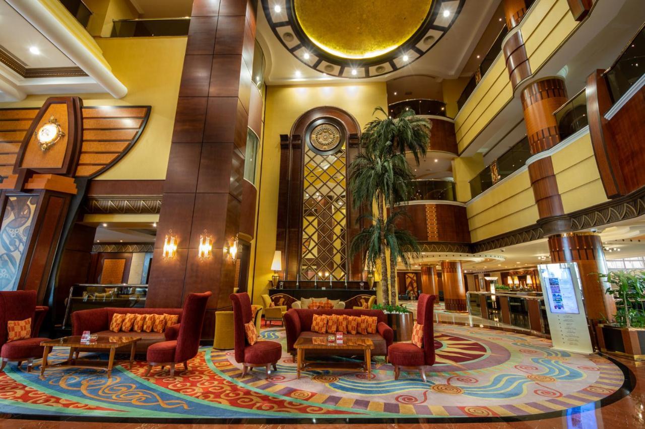 Al Raha Beach Resort & Spa Abu Dhabi Eksteriør bilde The photo shows an elegant lobby interior of a hotel. The space features tall ceilings with decorative lighting and large windows, allowing for natural light. There are several plush seating arrangements, including red armchairs and wooden tables, cr