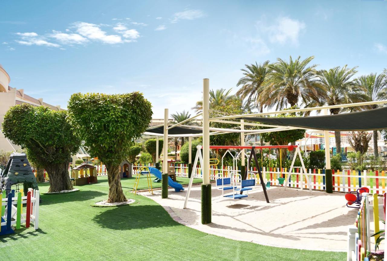 Al Raha Beach Resort & Spa Abu Dhabi Eksteriør bilde The photo shows a brightly lit outdoor playground surrounded by palm trees. The playground features a variety of play equipment, including swings, a slide, and a climbing structure. The ground is covered in lush green artificial grass, providing a so