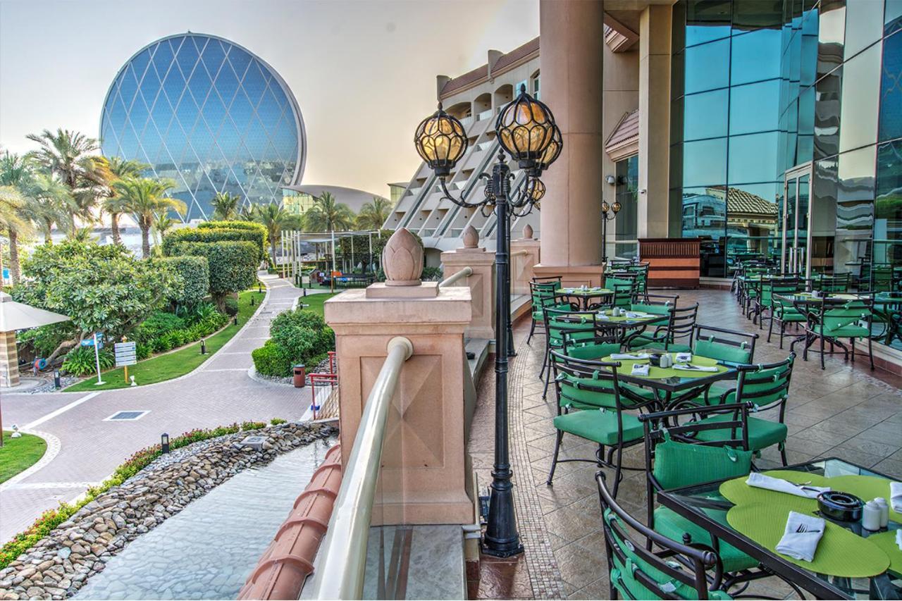 Al Raha Beach Resort & Spa Abu Dhabi Eksteriør bilde The photo depicts an outdoor dining area with green tables and chairs arranged on a patio. In the background, there is a large, distinctive building with a striking blue dome, surrounded by well-maintained landscaping, including trees and plants. The