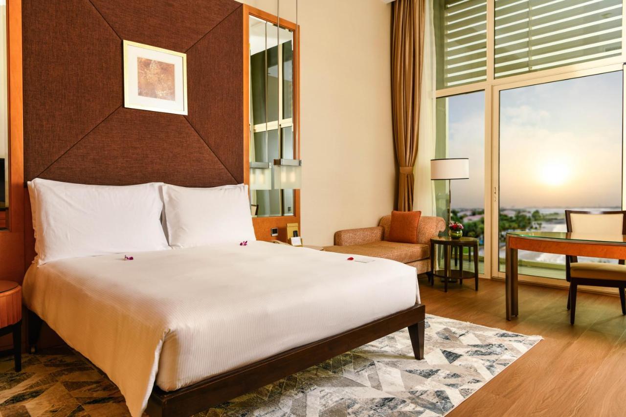 Al Raha Beach Resort & Spa Abu Dhabi Eksteriør bilde The photo shows a stylish hotel room featuring a large bed with white bedding, positioned against a textured wall. There’s a small seating area with a chair and a table, and a window with curtains that allows natural light to fill the space. The room