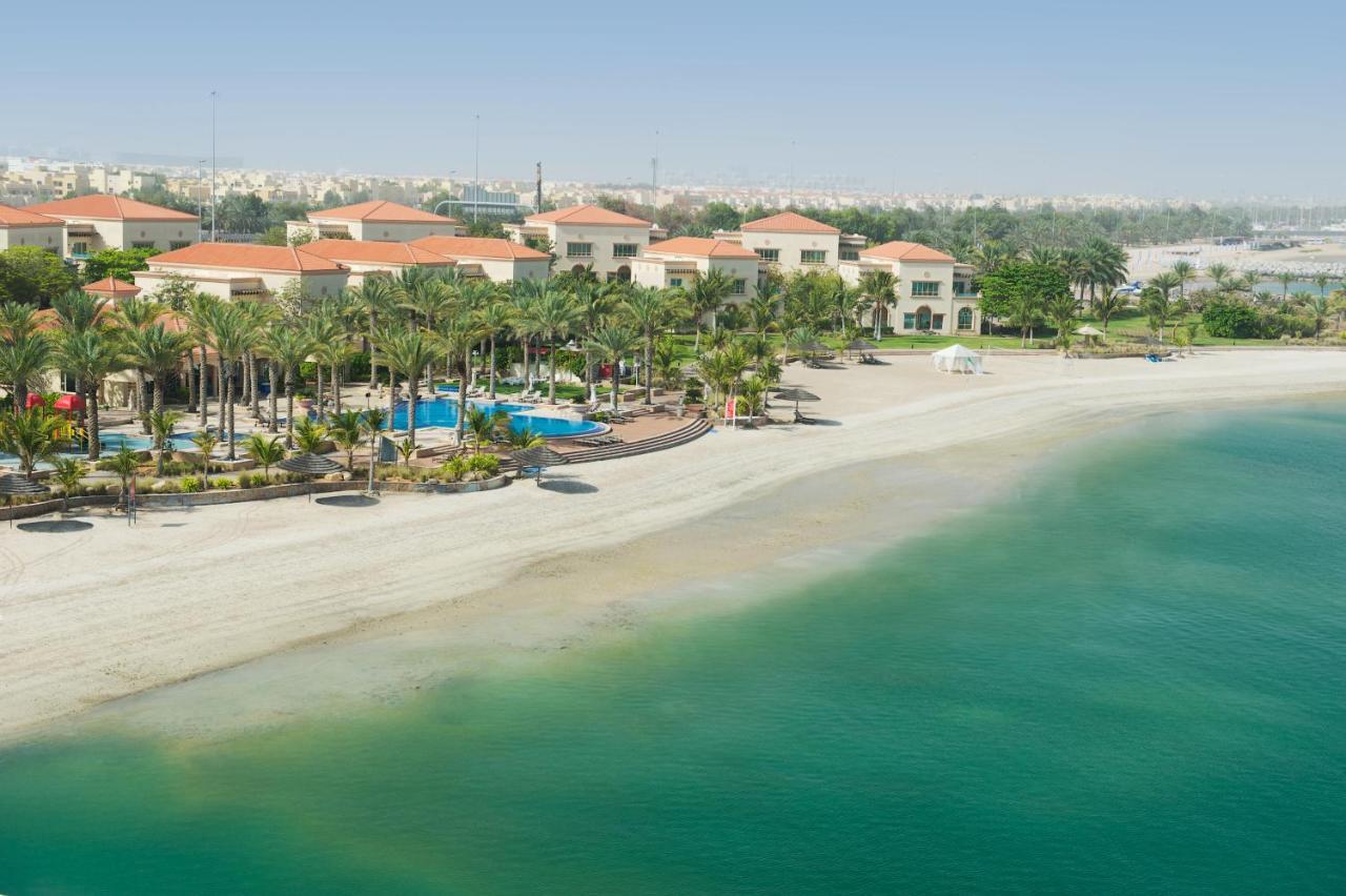 Al Raha Beach Resort & Spa Abu Dhabi Eksteriør bilde The photo depicts a serene beach scene with a clear turquoise sea and a sandy shoreline. In the background, there are several buildings featuring a Mediterranean architectural style with red-tiled roofs. The area is adorned with palm trees and outdoo