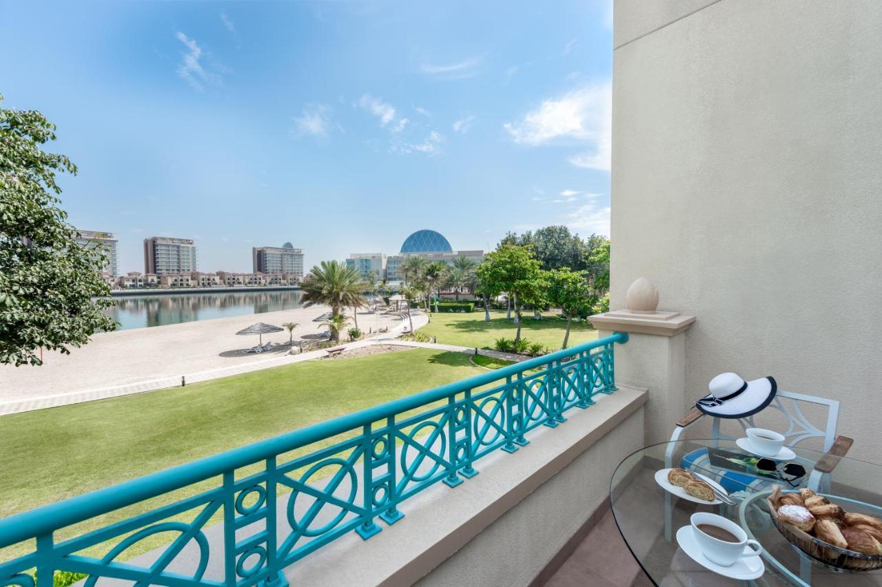 Al Raha Beach Resort & Spa Abu Dhabi Eksteriør bilde The photo shows a balcony view overlooking a scenic landscape. In the foreground, there is a glass table set with snacks or pastries and two cups. Beyond the balcony railing, there is a well-maintained green area with palm trees and a pathway. The ba