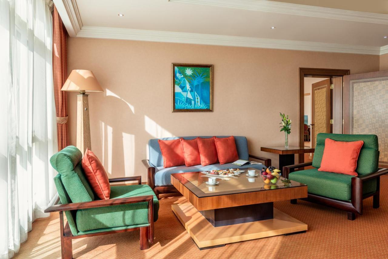 Al Raha Beach Resort & Spa Abu Dhabi Eksteriør bilde The photo shows a cozy living room with a warm ambiance. There are two sofas: one in green with red cushions and another in blue with red cushions, arranged around a wooden coffee table. On the table, there are some fruits and a small decorative item