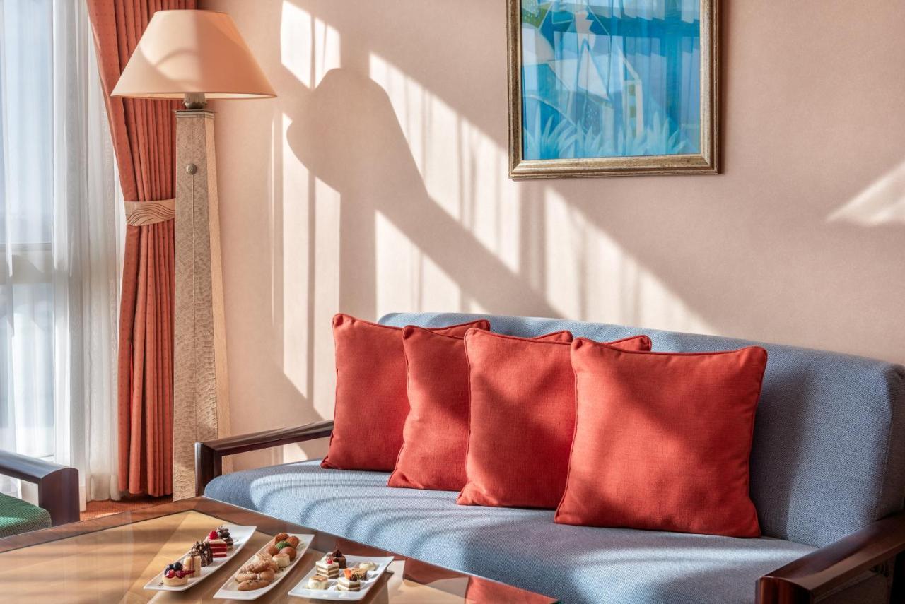 Al Raha Beach Resort & Spa Abu Dhabi Eksteriør bilde The photo shows a cozy interior setting, likely a sitting area. It features a light blue couch with four red cushions. In front of the couch, there is a glass table displaying an assortment of snacks or small dishes. The room has soft lighting, with 