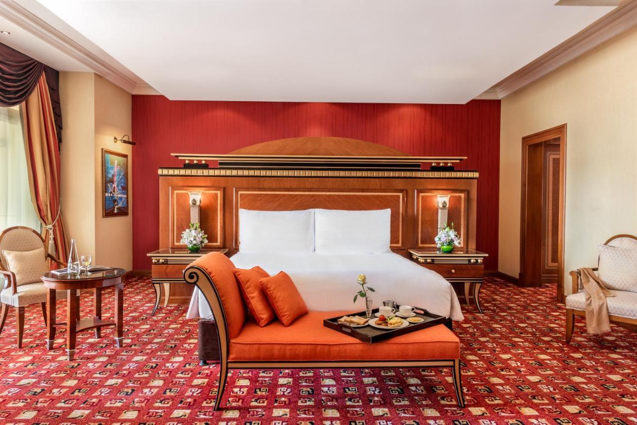 Al Raha Beach Resort & Spa Abu Dhabi Eksteriør bilde The photo shows an elegantly designed hotel room. The room features a large bed with white linens and a decorative headboard. The walls are painted in a warm red color, and there are side tables with lamps beside the bed. 

There is an orange sofa or