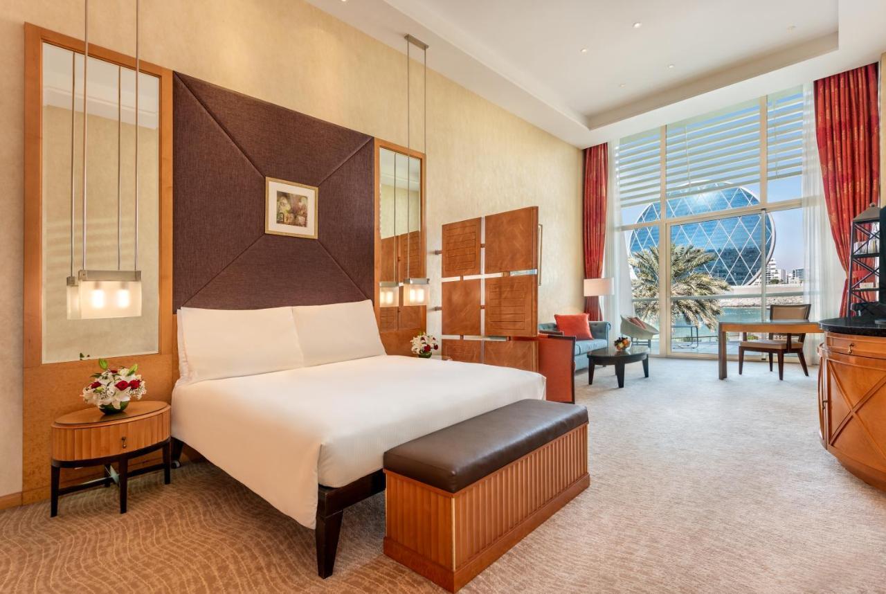 Al Raha Beach Resort & Spa Abu Dhabi Eksteriør bilde The photo shows a modern hotel room featuring a neatly made bed with a white comforter, situated against a stylish headboard with a geometric design. There are bedside tables on either side of the bed, each with a small flower arrangement. A comforta