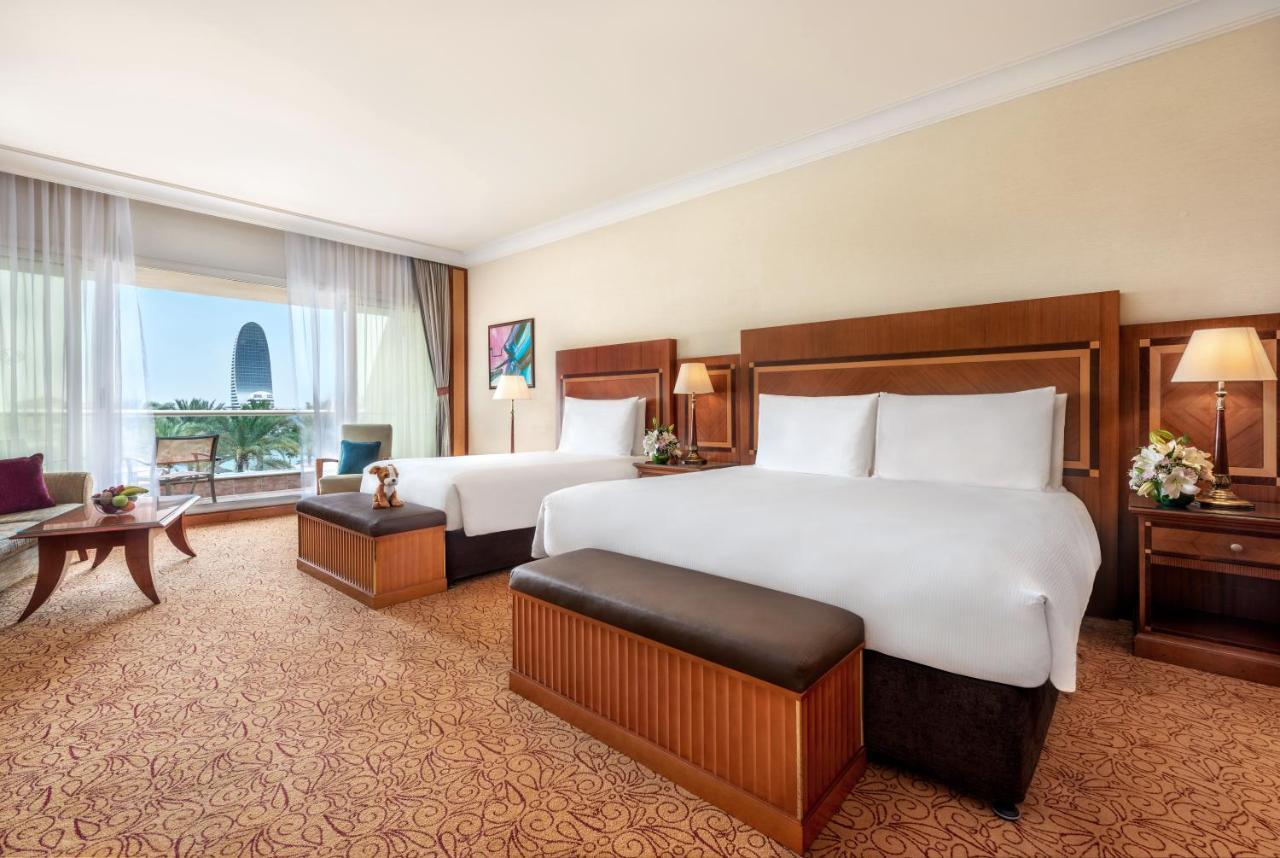 Al Raha Beach Resort & Spa Abu Dhabi Eksteriør bilde The photo shows a spacious hotel room featuring two large beds with white linens and dark brown upholstered headboards. There are matching benches at the foot of each bed. The room is decorated with a warm color palette and has a carpeted floor. To o