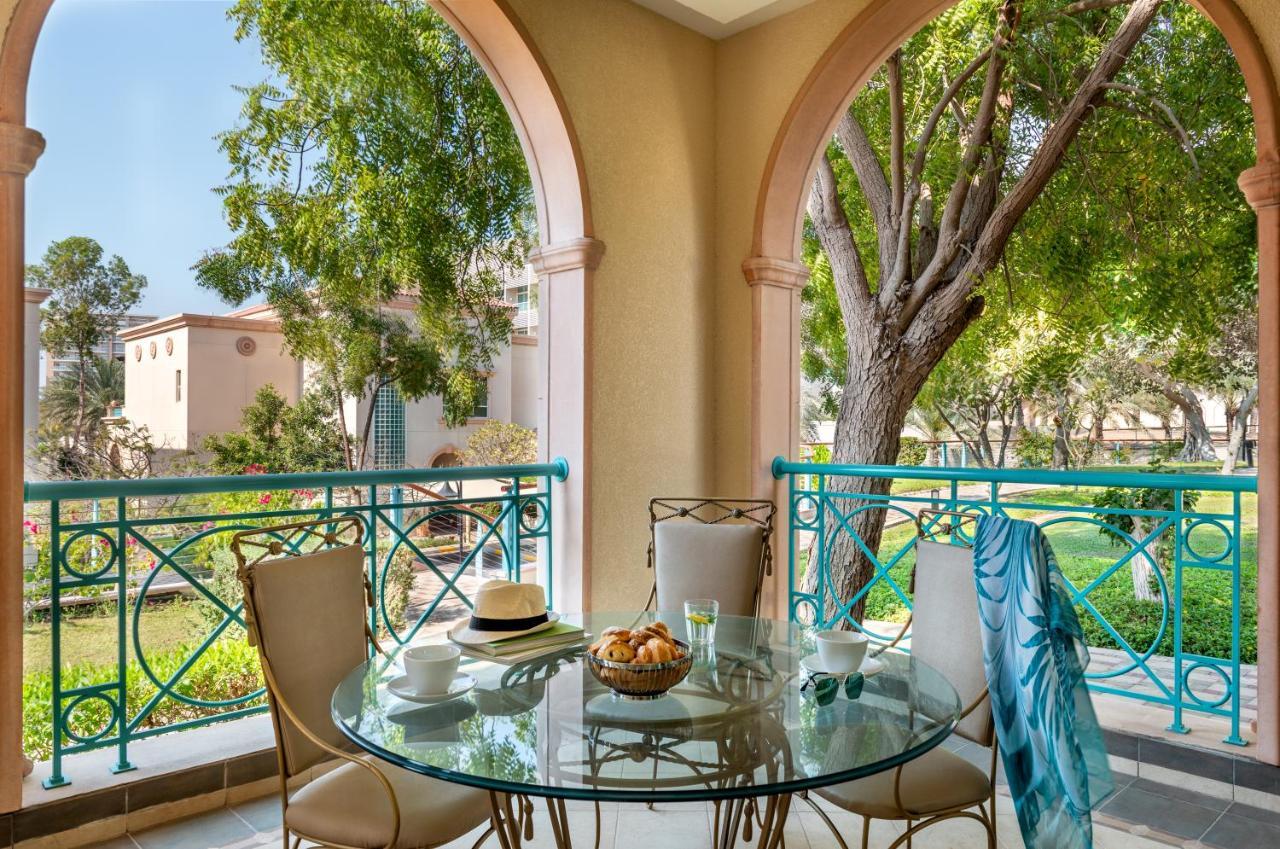Al Raha Beach Resort & Spa Abu Dhabi Eksteriør bilde The photo shows a serene outdoor balcony setting. There is a circular glass table with a bowl of fruit or snacks in the center, alongside a glass and a small plate. Surrounding the table are four elegant chairs. The railing is turquoise, framing a vi
