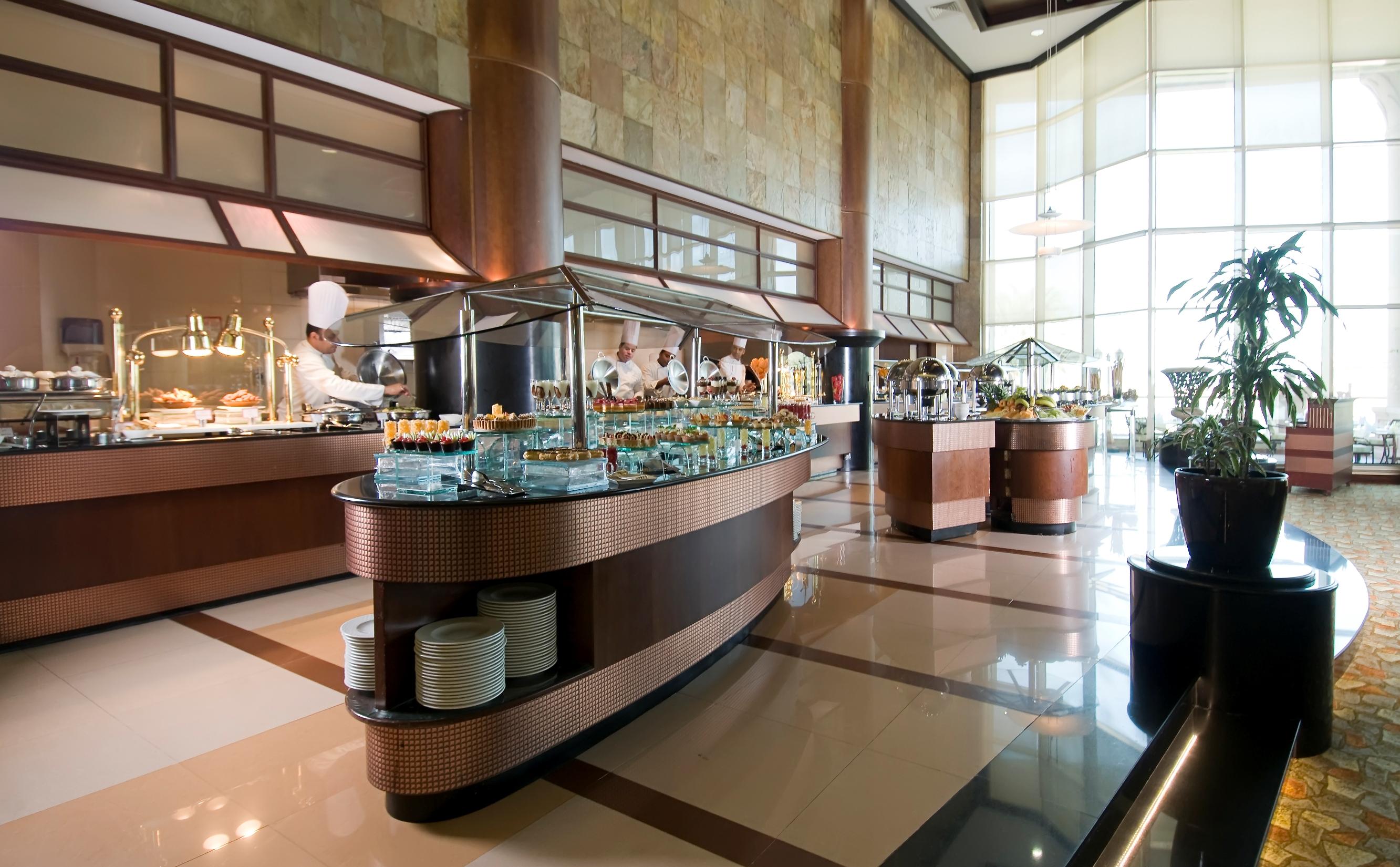 Al Raha Beach Resort & Spa Abu Dhabi Eksteriør bilde The photo shows a spacious buffet area in a restaurant or hotel. There are multiple food stations with glass displays showcasing a variety of dishes. Chefs can be seen in the background, preparing food. The decor features large windows allowing natur