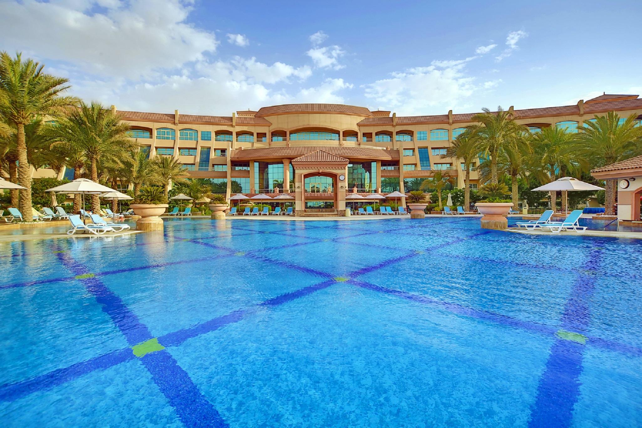 Al Raha Beach Resort & Spa Abu Dhabi Fasiliteter bilde The photo features a luxurious resort with a large swimming pool in the foreground. The pool is crystal clear and reflects the sky, bordered by lounge chairs and palm trees. In the background, the resort building showcases modern architecture with la