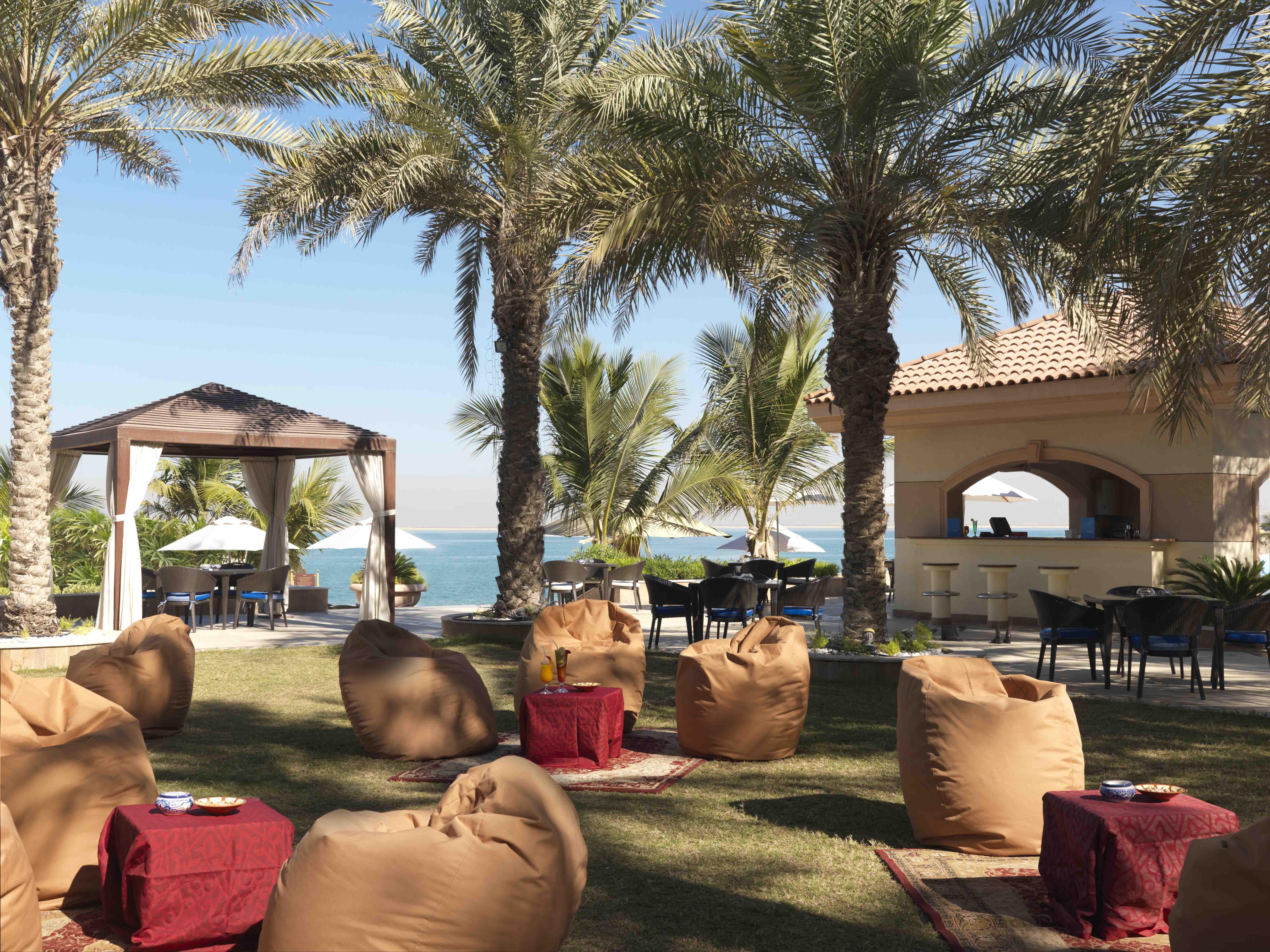 Al Raha Beach Resort & Spa Abu Dhabi Restaurant bilde The photo depicts a beautiful outdoor setting with several elements of relaxation and leisure. In the foreground, there are large, comfortable bean bag chairs arranged on a grassy area, accompanied by low tables. Surrounding this seating area are tal