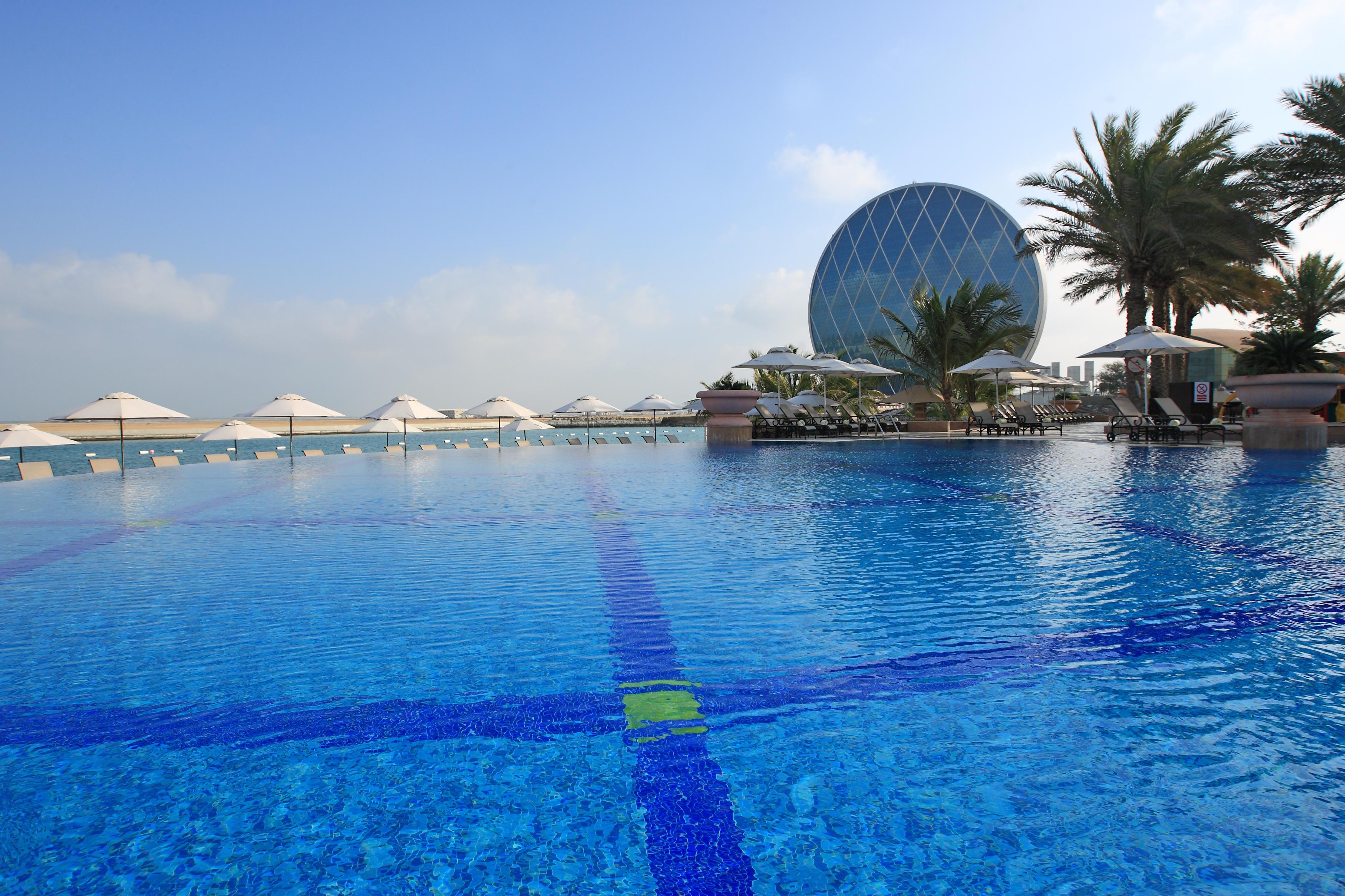 Al Raha Beach Resort & Spa Abu Dhabi Eksteriør bilde The photo features a large, serene swimming pool with clear, blue water reflecting the sky. Surrounding the pool are several lounge chairs and umbrellas, suggesting a relaxed, resort-like atmosphere. In the background, a distinctive building with a r