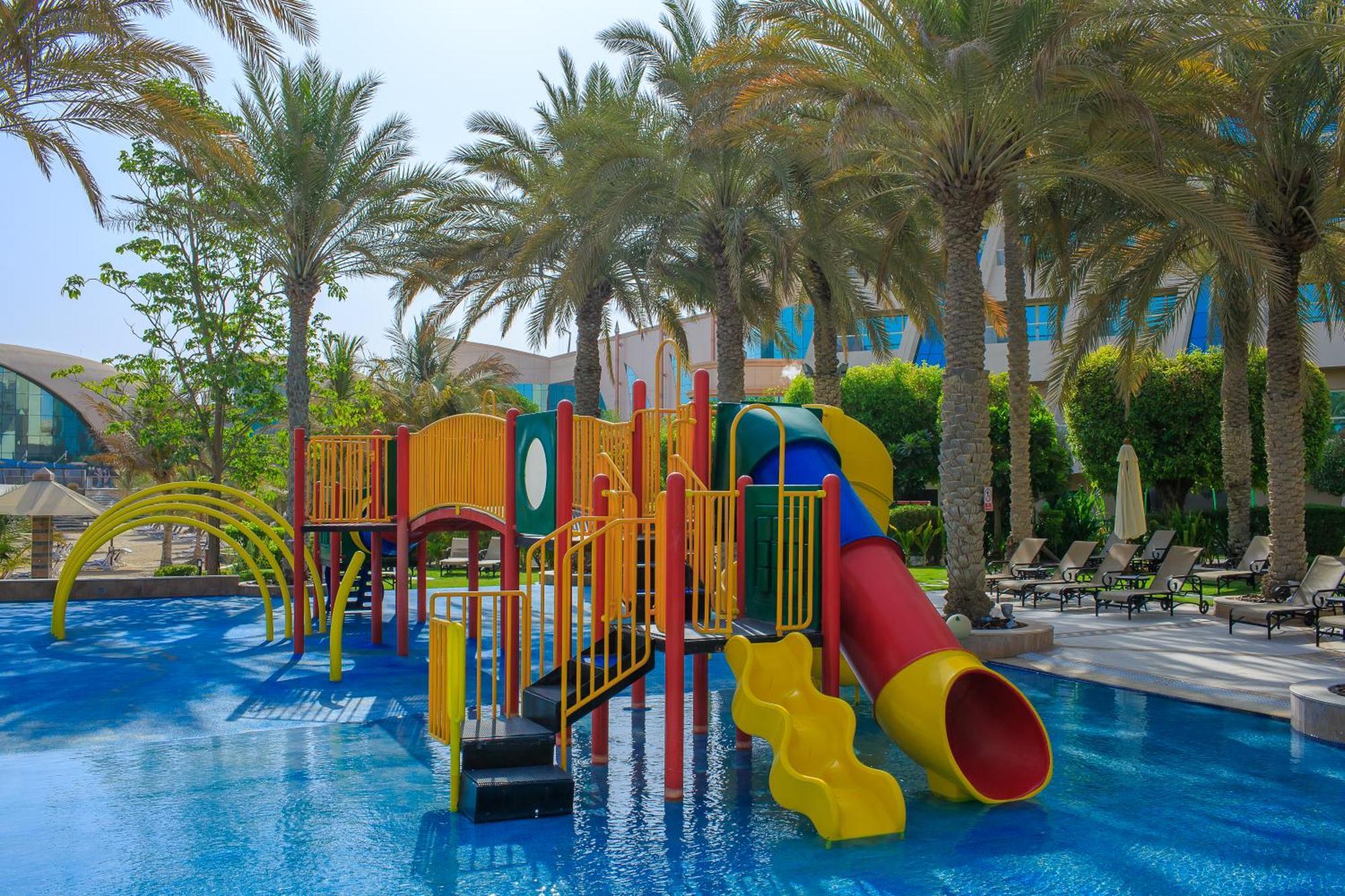 Al Raha Beach Resort & Spa Abu Dhabi Eksteriør bilde The photo shows a colorful water play area located in a pool setting. It features a variety of play structures, including slides in multiple colors, such as yellow, red, and blue. Surrounding the play structure are palm trees, creating a tropical atm