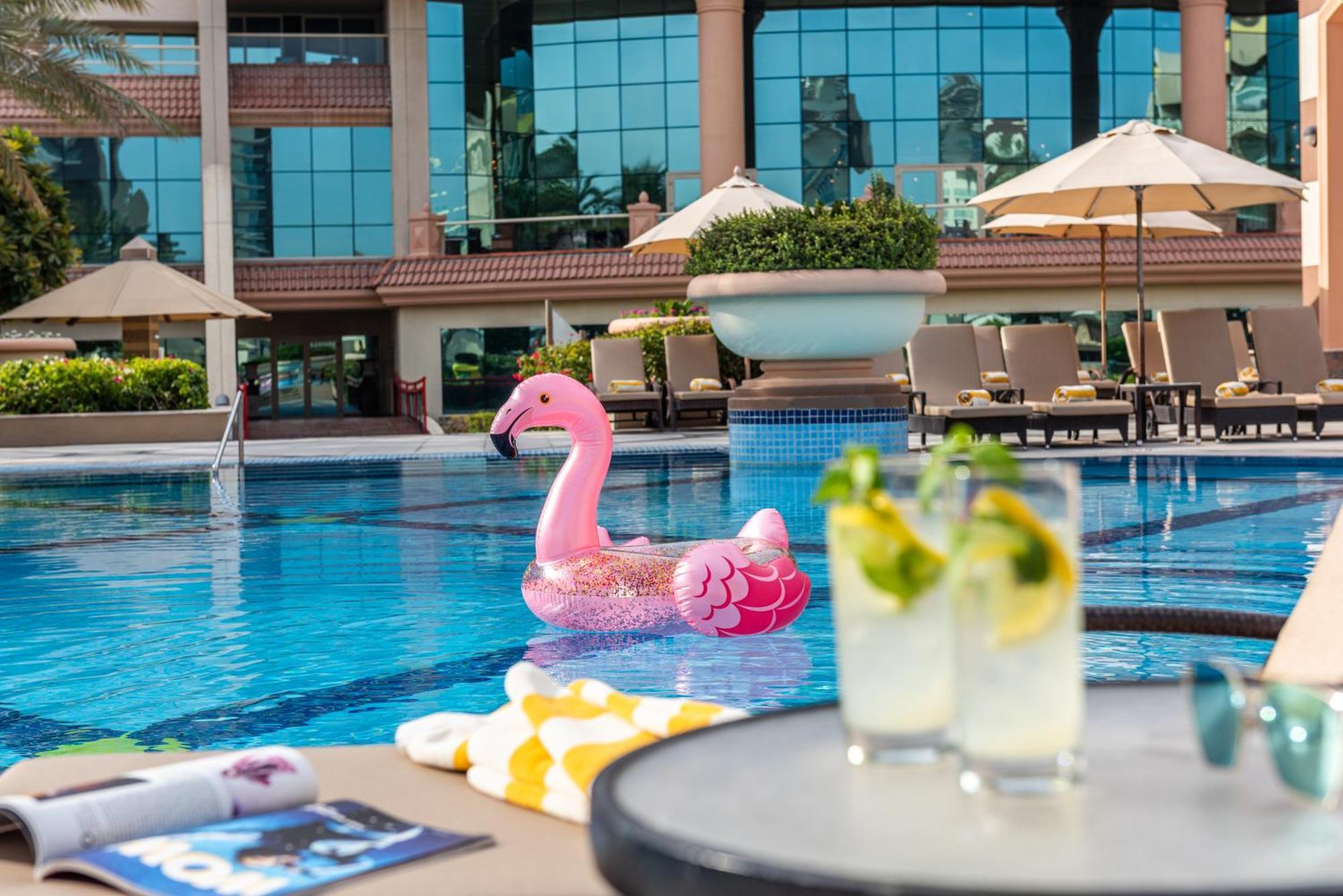 Al Raha Beach Resort & Spa Abu Dhabi Eksteriør bilde The photo depicts a bright and inviting outdoor swimming pool surrounded by a luxurious setting. In the pool, there is a large pink flamingo float, adding a fun and whimsical touch. To the side, a table is set up with two refreshing drinks, likely co