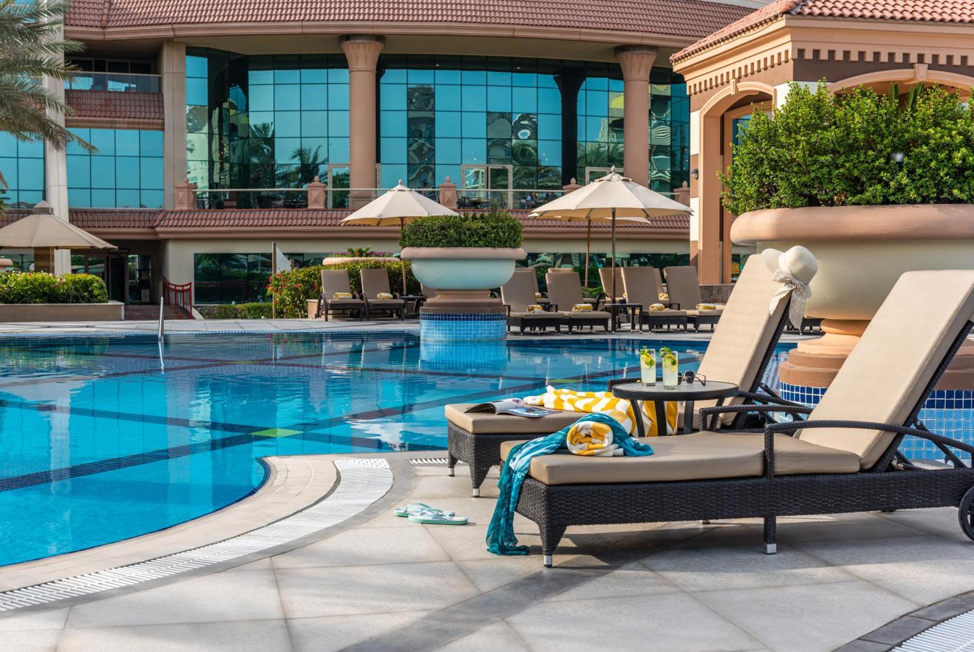 Al Raha Beach Resort & Spa Abu Dhabi Eksteriør bilde The photo depicts a luxurious outdoor swimming pool area. In the foreground, there are two lounge chairs with cushions, positioned beside the pool. A small table between them holds drinks and snacks, suggesting a relaxing atmosphere. The pool itself 