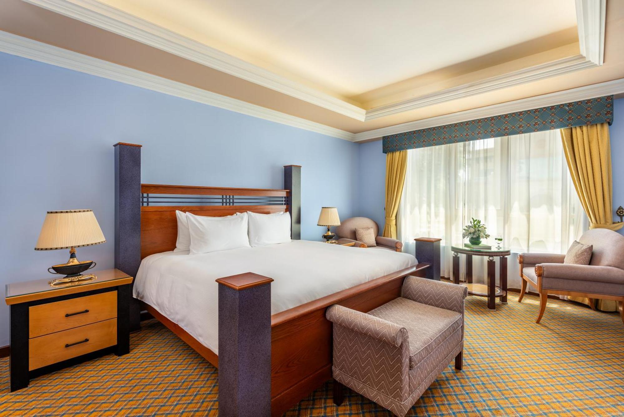 Al Raha Beach Resort & Spa Abu Dhabi Eksteriør bilde The photo shows a well-decorated hotel room. It features a large bed with a wooden frame, neatly made with white bedding. On either side of the bed are bedside tables with lamps. There is a comfortable chair next to a window that has sheer curtains a