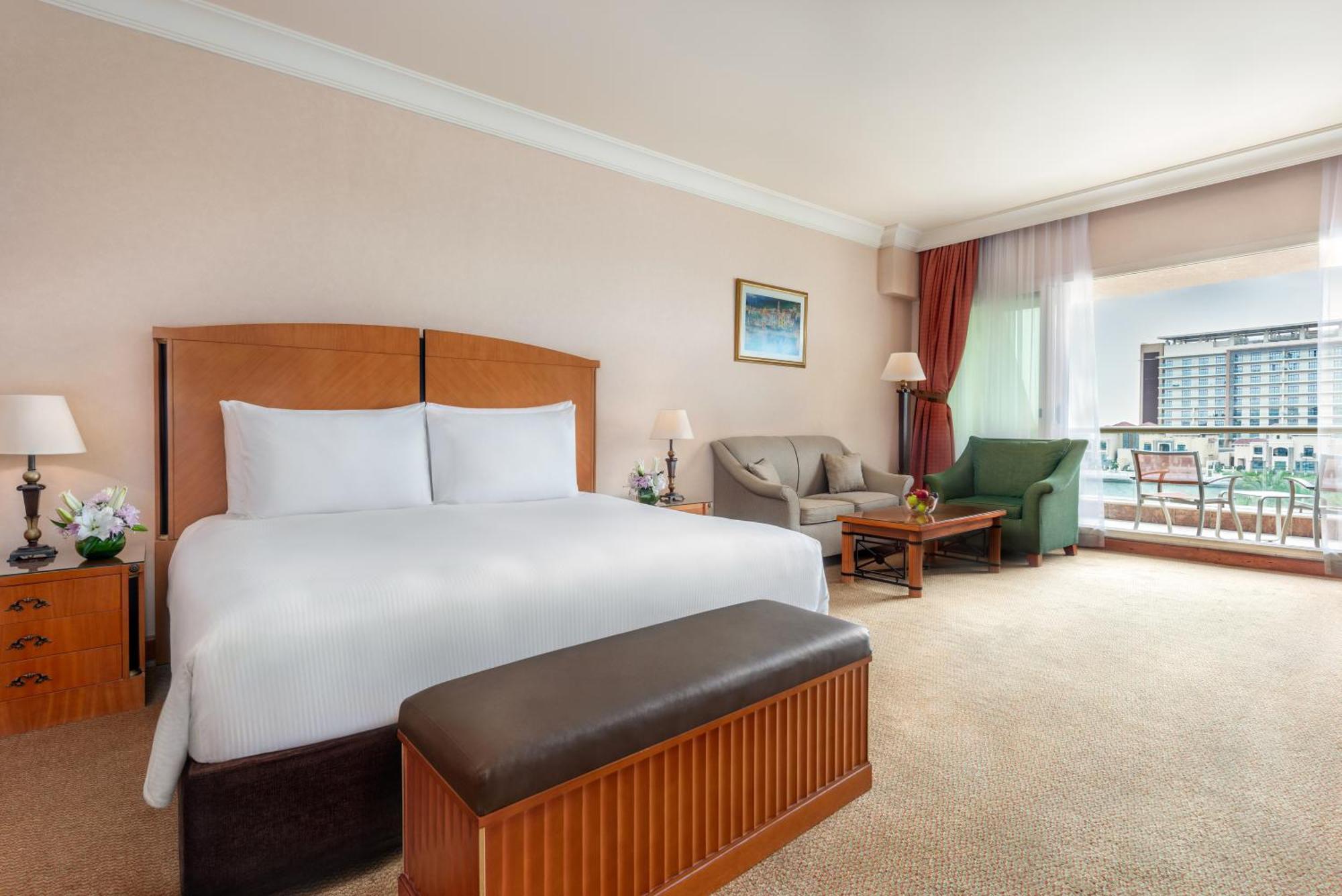 Al Raha Beach Resort & Spa Abu Dhabi Eksteriør bilde The photo shows a well-furnished hotel room featuring a large bed with white linens and a wooden headboard. There's a brown bench at the foot of the bed. To one side, a sitting area includes a sofa and a coffee table, complemented by two lamps. Large