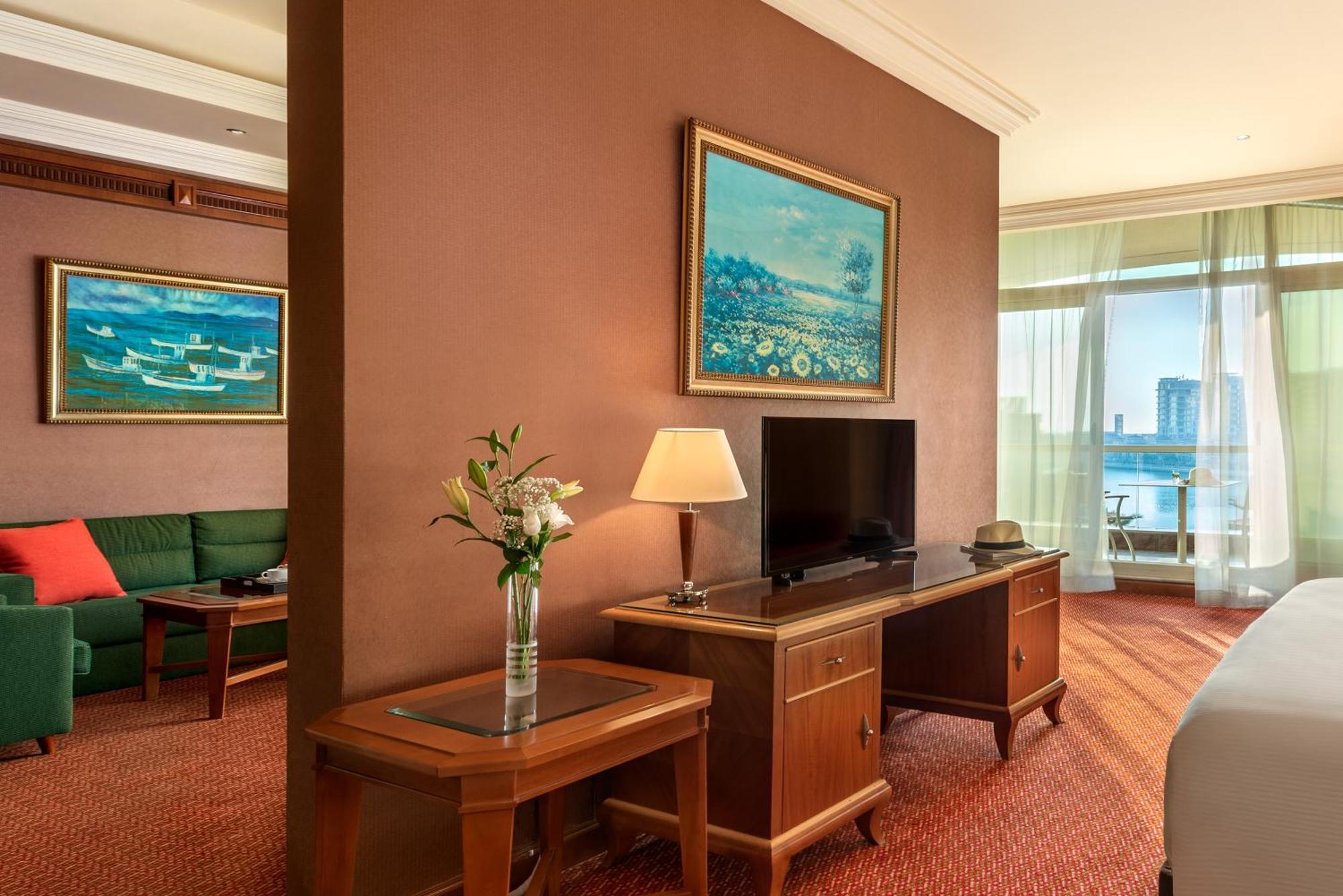 Al Raha Beach Resort & Spa Abu Dhabi Rom bilde The photo shows a hotel room with a warm, inviting atmosphere. There's a large window that provides natural light and a view, likely overlooking a cityscape or water. On one wall, there's a piece of artwork featuring a landscape, and nearby, a small 