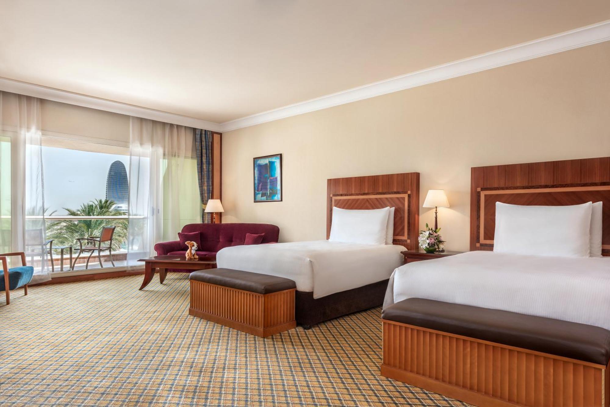 Al Raha Beach Resort & Spa Abu Dhabi Rom bilde The photo shows a hotel room featuring two twin beds with wooden headboards. The room has a light, neutral color scheme, with a large window that provides natural light and a view of the outdoors. There is a small seating area with a purple sofa and 