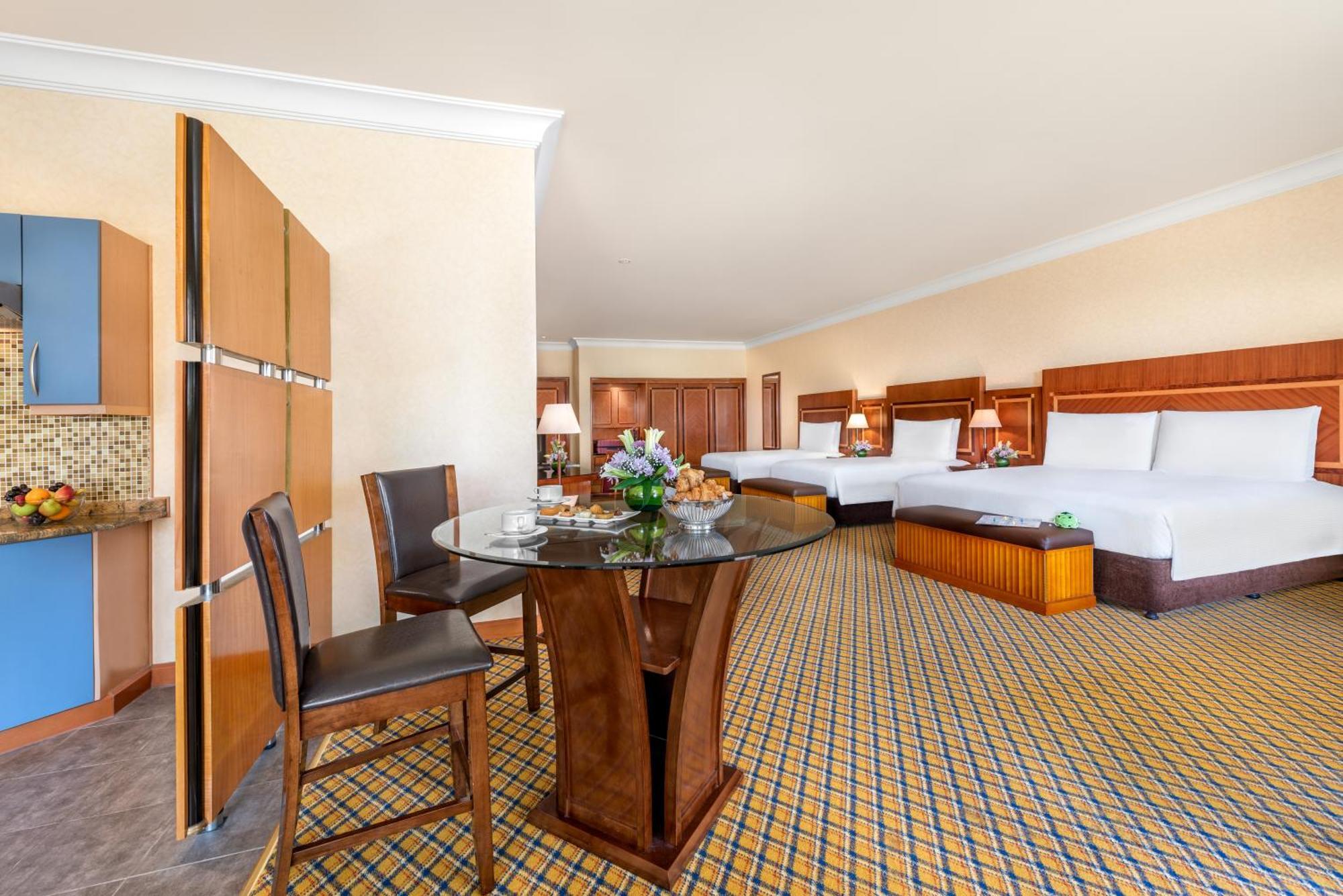 Al Raha Beach Resort & Spa Abu Dhabi Rom bilde The photo displays a spacious hotel room. It features two large beds with white bedding, a circular glass dining table with a decorative floral centerpiece, and a couple of chairs around it. There is a wooden cabinet along one wall, and the room has 