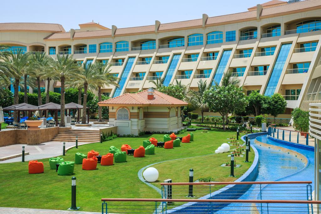 Al Raha Beach Resort & Spa Abu Dhabi Eksteriør bilde The photo depicts a modern hotel complex with multiple stories and large windows. In the foreground, there is a well-maintained lawn featuring colorful lounge chairs in shades of orange and green. A pathway runs alongside a blue water feature, creati