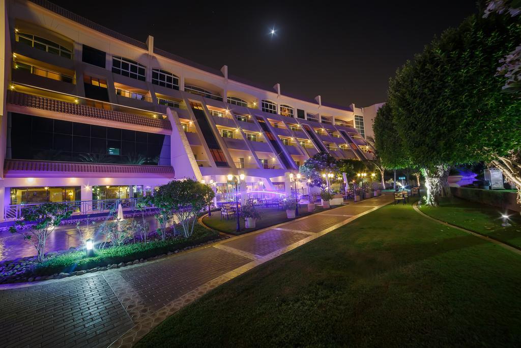 Al Raha Beach Resort & Spa Abu Dhabi Eksteriør bilde The photo shows a modern building illuminated at night, with a vibrant purple hue. The structure has multiple floors, each with balconies overlooking a landscaped area. In the foreground, there is a pathway lined with well-manicured grass and shrubs,