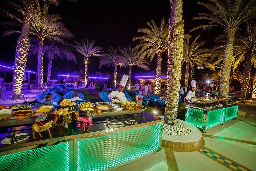 Al Raha Beach Resort & Spa Abu Dhabi Eksteriør bilde The photo depicts an outdoor dining area at night, featuring several chefs in white uniforms and hats working at a food station. The setting is illuminated by colorful lights, with glowing green accents on the serving tables. Surrounding the area are